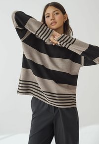 Pull rayé noir et beige, avec de larges rayures horizontales, des poignets côtelés et une coupe décontractée, assorti à un pantalon sombre.