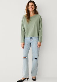 Hellgrünes Langarmoberteil, kombiniert mit hellblauen, verwaschenen Jeans und schwarzen spitz zulaufenden Heels, bietet einen lockeren Sitz und einen weichen Stoff.