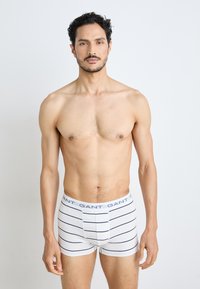 Boxer briefs blancs avec des rayures horizontales navy, dotés d'une taille de marque. Le modèle a un physique tonique, sur un fond uni.