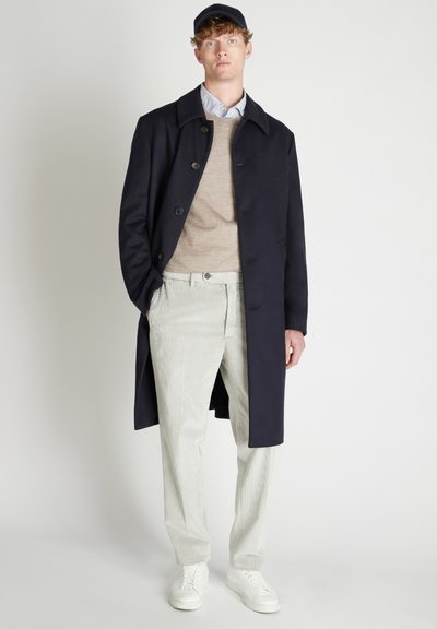 Giovane uomo che indossa un soprabito blu marino, un maglione beige, una camicia a righe blu, pantaloni grigi chiari, sneakers bianche e un cappellino blu marino, in piedi contro uno sfondo semplice.