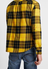 Żółto-granatowa koszula flanelowa w kratę z mankietami na guziki, zestawiona z niebieskimi dżinsami z ozdobnymi kieszeniami z tyłu i widocznymi przeszyciami.