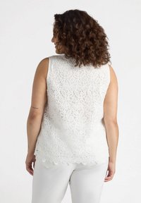 White lace sleeveless top with floral pattern, scalloped hem, and sheer texture. Worn over light-colored bottoms, viewed from the back.