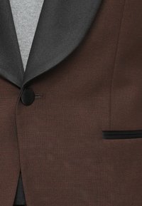 Sacou texturat maro cu rever din satin gri închis, un buton negru și un buzunar pe piept cu margine neagră, purtat peste un top din tricot gri deschis.