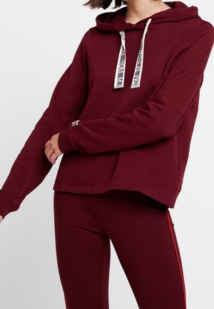 Vrouw die een bordeauxrode hoodie draagt met witte trekkoorden en bijpassende bordeauxrode leggings met een rode zijstreep.