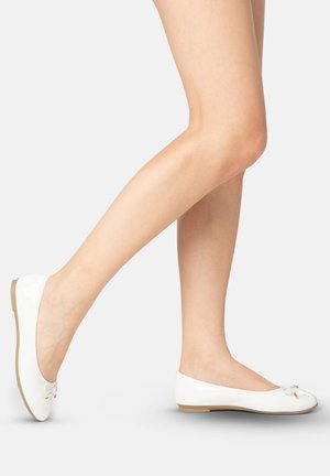 Piernas que llevan bailarinas blancas con pequeños lazos, un pie levantado por detrás, sobre un fondo claro y liso.