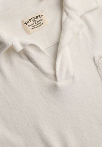 Vêtement en tissu texturé blanc avec col ouvert et étiquette beige indiquant "Superdry Co Craft & Detail Trade Mark British Design."