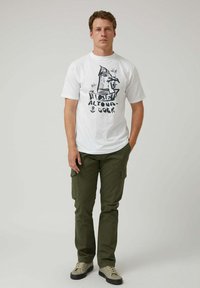Camiseta blanca de algodón con un diseño gráfico de un barco y texto; combinada con pantalones cargo verde oliva; zapatillas deportivas en tonos neutros.