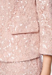 Blush-pink Spitzenjacke mit floralen Mustern, verziert mit Pailletten. Enthält eine Fronttasche mit gewellten Kanten und strukturierter Textur.