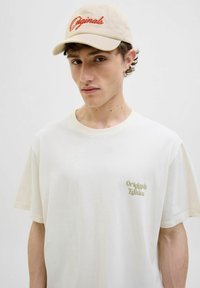 Beige baseball cap med rød "Originals" brodering, parret med en off-white oversized t-shirt med subtil "Originals Edition" tekst i grøn.