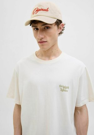 Beige baseball cap med rød "Originals" brodering, parret med en off-white oversized t-shirt med subtil "Originals Edition" tekst i grøn.