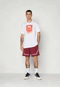Witte katoenen t-shirt met een kleurrijke grafische print. Kastanjebruine shorts met zijaccenten en witte strepen. Zwart-witte sportieve schoenen.