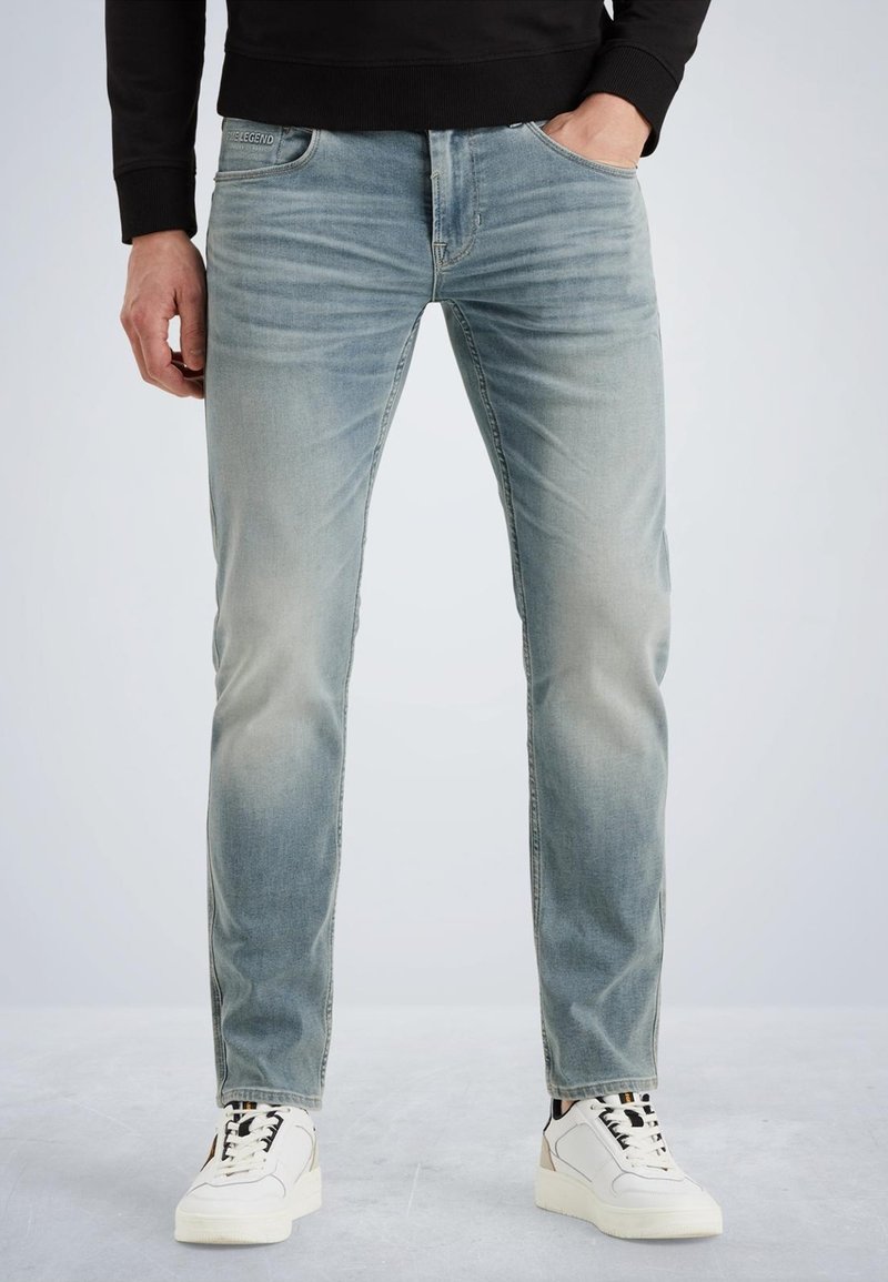 Jean en denim bleu clair avec une coupe fuselée, texture délavée, design à cinq poches et un détail de marque discret sur la ceinture. Associé à des baskets blanches.