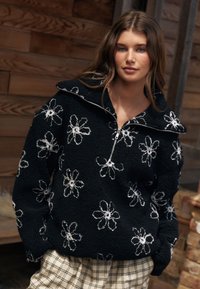 Sort fleece-pullover med høj krave og sølv lynlås, med hvide blomstermønstre over det hele. Struktureret stof og afslappet pasform.
