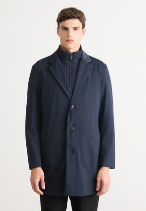 Man wearing a navy blue buttoned coat over a zipped high-collar inner layer against a plain white background.