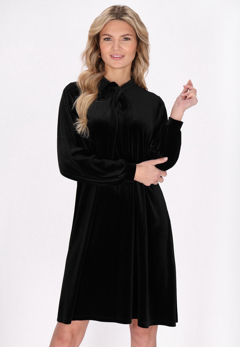 Schwarzes Samtkleid mit langen Ärmeln, einem Schleifenhalsauschnitt und einer schwingenden Silhouette. Der Stoff wirkt weich und glatt.