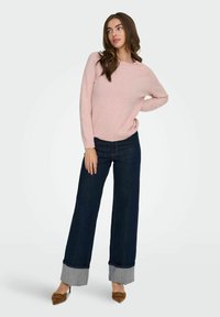 Maglione rosa chiaro con una vestibilità comoda, abbinato a jeans a gamba larga blu scuro con un risvolto a contrasto. Tacchi marroni completano l'outfit.