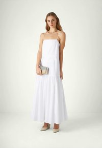 Νεαρή γυναίκα σε λευκό άφορο μάξι φόρεμα, κρατώντας μια ασημένια τσάντα, φορώντας μπεζ ψηλοτάκουνα και χρυσά κοσμήματα, στέκεται μπροστά σε λευκό φόντο.