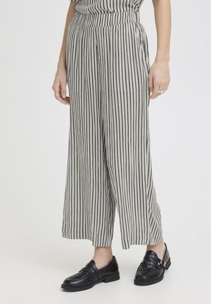 Donna che indossa pantaloni beige a gamba larga con strisce verticali nere e mocassini slip-on in pelle nera su sfondo bianco semplice.