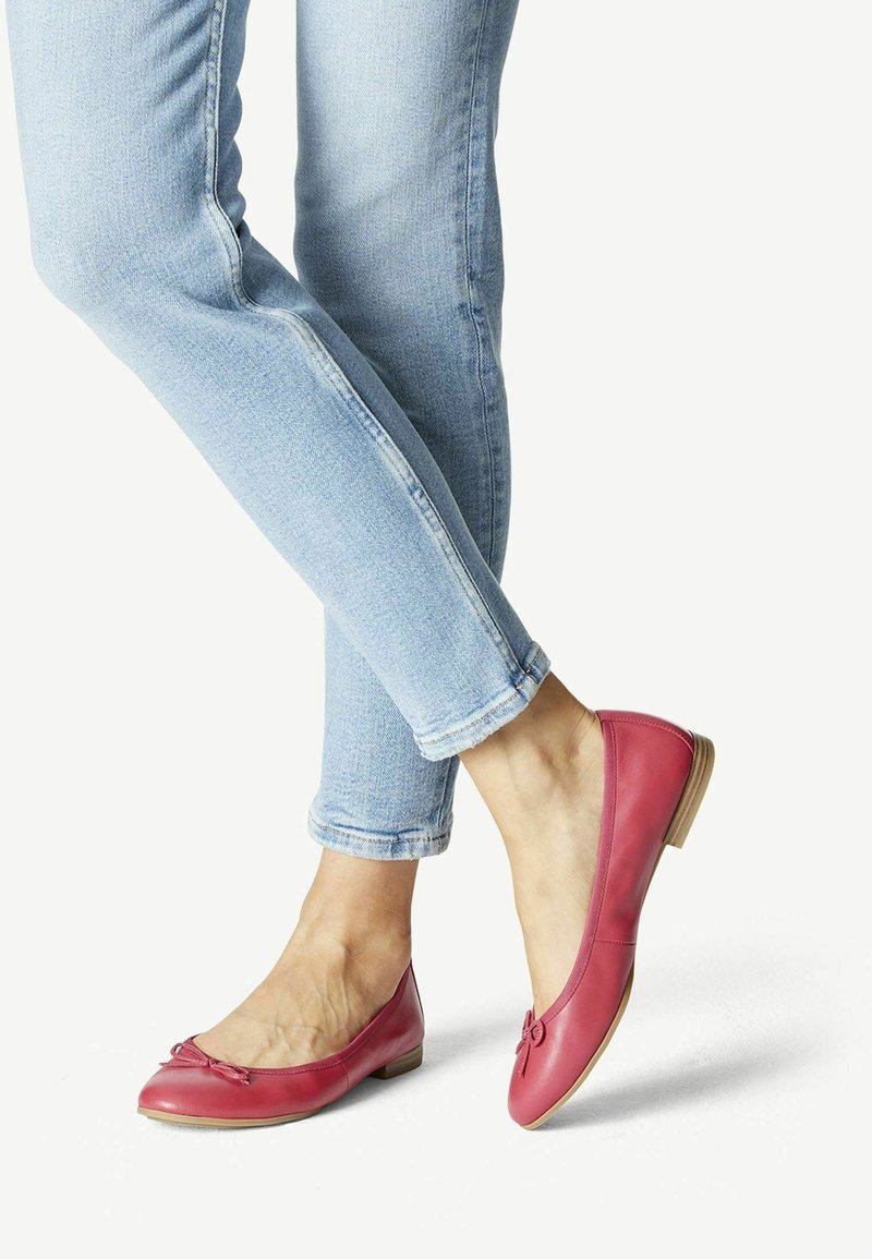 Jambes croisées portant un jean skinny bleu clair et des ballerines en cuir rouge avec de petits nœuds, sur fond blanc.