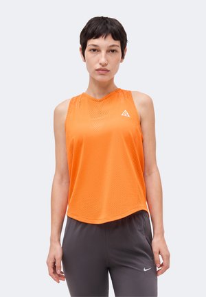 Kobieta z krótkimi ciemnymi włosami ubrana w pomarańczowy siateczkowy top bez rękawów oraz ciemnoszare spodnie sportowe na jednolitym jasnym tle.