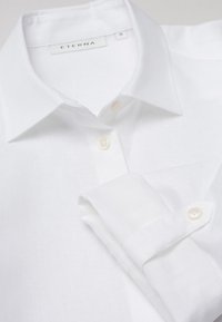 Camicia bianca in lino con colletto a bottoni, caratterizzata da una sottile texture. Include bottoni chiari e un dettaglio con il manico piegato. Marca: ETERNA.