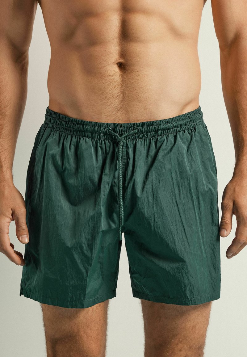Shorts de bain verts en tissu léger et froissé, avec une taille élastique et un cordon de serrage. Longueur mi-cuisse, sans motifs visibles.