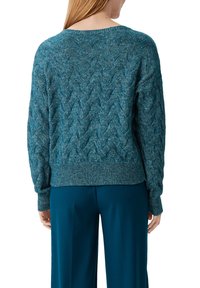Suéter de punto trenzado en tono verde azulado con cuello redondo, puños y dobladillo acanalados. Textura de punto grueso, llevado sobre pantalones holgados.