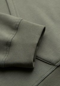 Lähedalt vaadatuna oliiviroheline spordipusa varrukas, millel on soonikkoes mansett ja detailsed õmblused siledal kangal.