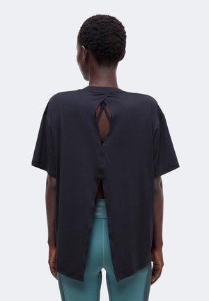 Personne aux cheveux courts et bouclés portant un haut noir ample à manches courtes avec une fente basse dans le dos et un pantalon ajusté bleu sarcelle, vue de dos.