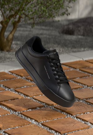 Svart läder sneaker från Tommy Hilfiger med snören, svävande över träplattor och grus utomhus nära ett träd.