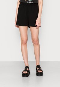 Shorts noirs légers avec une taille élastique, présentant une coupe décontractée. Associés à des sandales noires épaisses avec de larges brides et des semelles compensées.