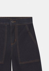 Donkerblauwe denim broek met opvallende oranje stiksels, voorkruiszakken en een knoopsluiting bij de taille.