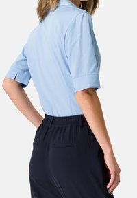 Hellblaue kurzärmelige Bluse mit Rückenfalte, kombiniert mit marineblauen Hosen mit elastischem Bund und zwei Gesäßtaschen.