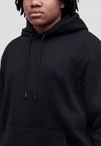 Person, die einen schlichten schwarzen Hoodie mit Kordeln trägt, vom Hals bis zur Mitte des Oberkörpers vor neutralem Hintergrund gezeigt.
