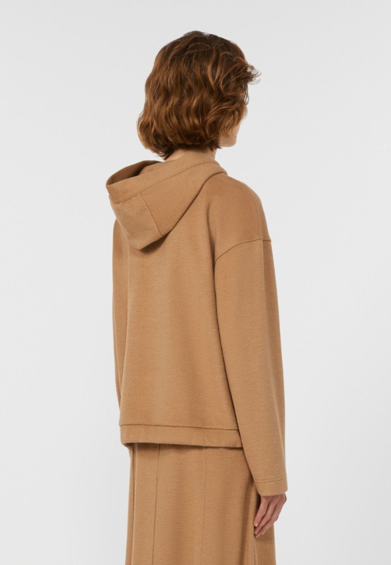 Person mit kurzen, lockigen braunen Haaren, die eine lockere, camel-farbene Kapuzenjacke und einen passenden Rock trägt, mit dem Rücken zu einem weißen Hintergrund gewandt.