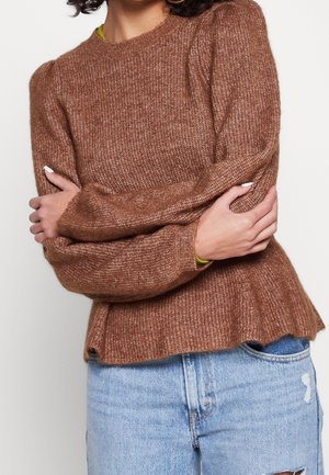Pull en maille marron avec des manches bouffantes, col rond et ourlet côtelé. Associé à un short en jean déchiré, mettant en valeur un look superposé.