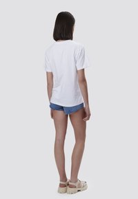 Witte katoenen t-shirt weergegeven vanuit de achterkant, gecombineerd met blauwe denim shorts en beige chunky sandalen, op een neutrale achtergrond.