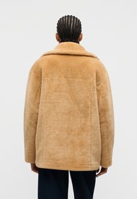 Άτομο με πλεξούδες που φοράει μπεζ τζάκετ με επένδυση από shearling και σκούρο παντελόνι, στέκεται γυρισμένο προς τα πίσω μπροστά σε απλό φωτεινό φόντο.