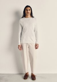 Hombre con cabello largo y bigote de pie contra una pared beige, vistiendo una camisa de manga larga gris claro, pantalones crema y zapatos de cuero marrón.