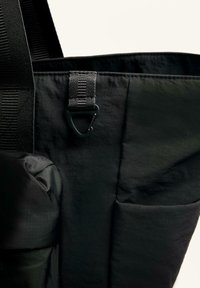 Schwarze Tragetasche aus strukturiertem Nylon, mit einem Clip-Verschluss, verstellbarem Tragegurt und einer Seitentasche für zusätzlichen Komfort.