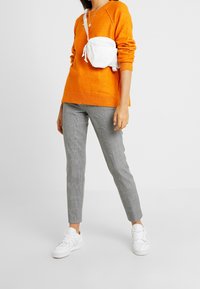 Oranžový pletený svetr, šedé kostkované kalhoty a bílé tenisky. Malá bílá crossbody kabelka spočívá na rameni. Čistý, ležérní vzhled.