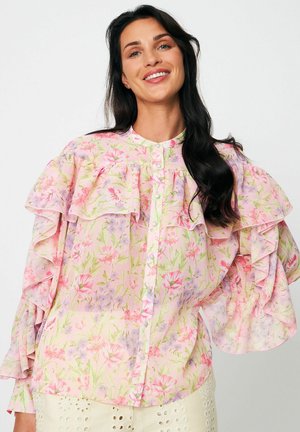 Naeratav naine pikkade tumedate juustega, kes kannab pastellsete lillemustriga pluusi, millel on volangid õlgadel ja varrukatel, valge tausta ees.