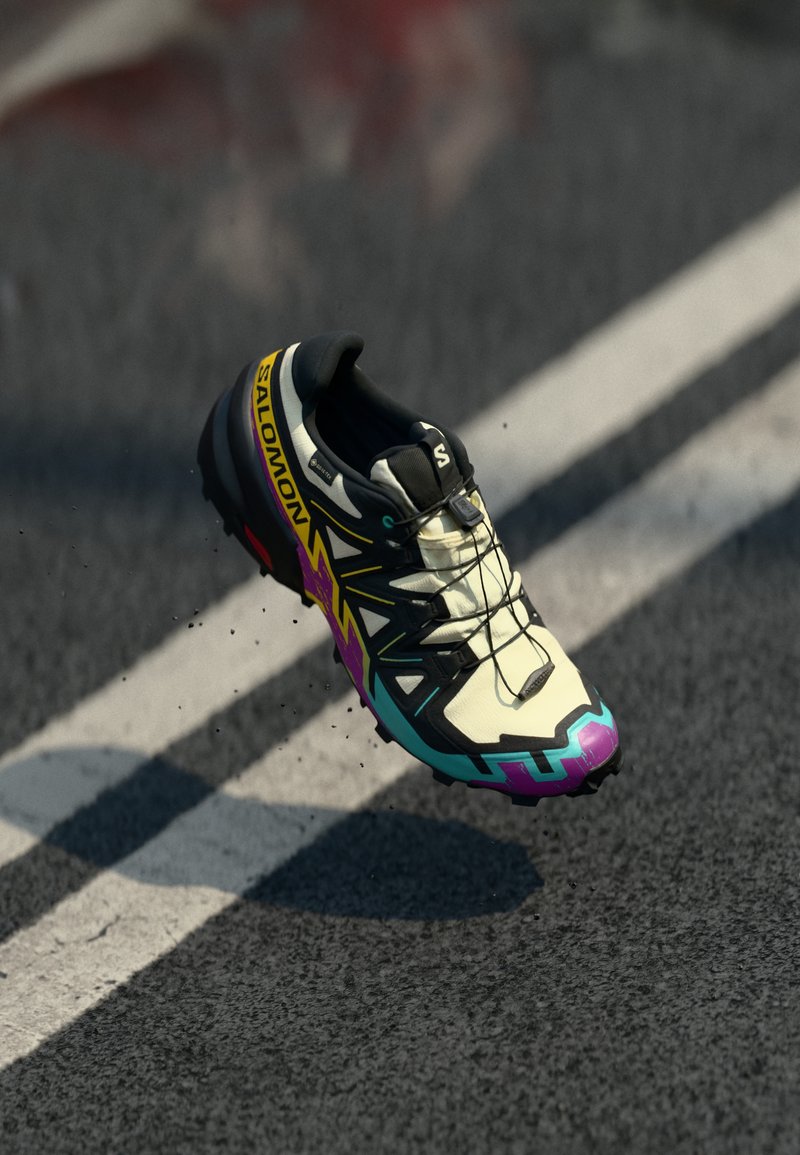 Chaussure de trail running avec un dessus blanc, des accents noirs, des motifs graphiques colorés et une semelle texturée pour l'adhérence. Logo de la marque visible.