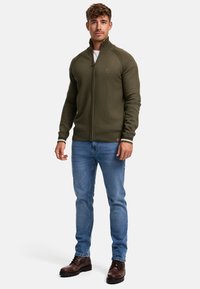 Oliven grønn zip-up genser med ribbestrikkede mansjetter og kant, med et teksturert strikkemønster og en liten logo på brystet. Brukt med blå jeans og brune støvler.