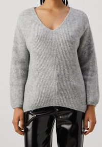 Grå stickad tröja med V-ringning, ribbad textur och långa ärmar. Bärs med glänsande svarta läderbyxor. Diskreta detaljer genom hela plagget.