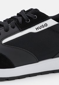 Chaussure de sport noire en mesh et suède ; comporte des accents blancs, une surface texturée, des lacets noirs et un logo de marque.