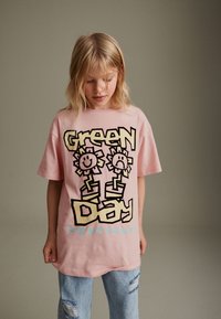 T-shirt di cotone rosa con il testo "Green Day" in nero e giallo e due fiori in stile cartone animato, abbinata a jeans chiari strappati.