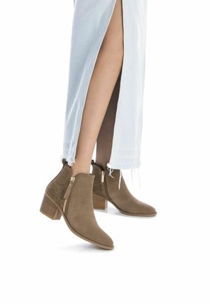 Botas de ante marrón hasta el tobillo con paneles laterales perforados, cierres con cremallera y tacones de bloque de 2.5 pulgadas, combinadas con una falda de mezclilla clara.