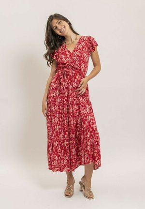 Femme souriante, portant une robe portefeuille fleurie rouge et rose avec des manches volantées et des sandales à talons hauts dorées, posant devant un fond uni.