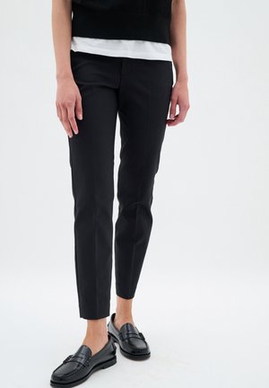 Person trägt schmale schwarze Hose und schwarze Lederloafers, steht vor einem einfarbigen hellen Hintergrund mit entspannten Händen seitlich am Körper.